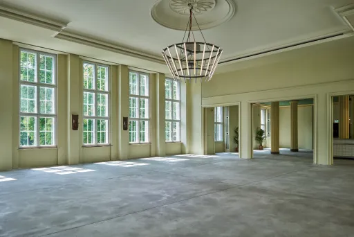 Großer, leerer Saal mit hohen Fenstern und Kronleuchter, viel Tageslicht.