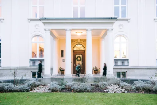 Weiße Villa mit Säulen, beleuchtetem Eingang und drei Personen im Vorgarten.