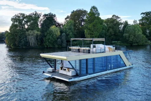 Modernes Hausboot mit Glasfront auf einem Fluss, umgeben von Bäumen.