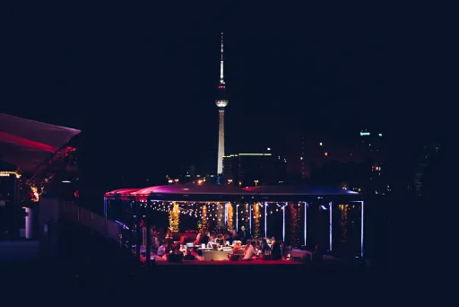 Beleuchtete Dachterrasse mit Menschen, im Hintergrund der Berliner Fernsehturm bei Nacht.