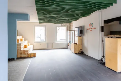 Heller Veranstaltungsraum mit großen Fenstern, modularer Holztribüne und flexibler Technik. An der Decke wellenförmige Elemente, an der Wand das Logo der Cradle to Cradle NGO. Offene Fläche für Workshops und Events.