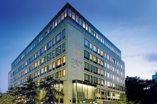 Modernes Hotelgebäude bei Dämmerung mit beleuchteten Fenstern und Bäumen davor.