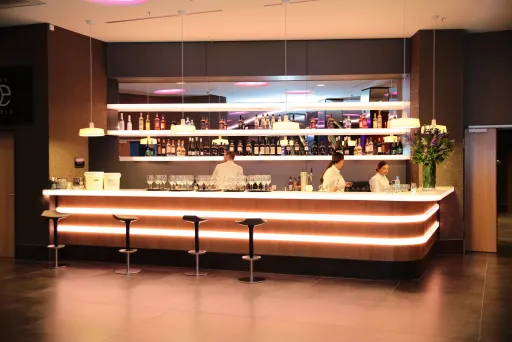 Moderne Bar mit drei Barhockern, zwei Barkeepern und beleuchteten Regalen im Hintergrund.
