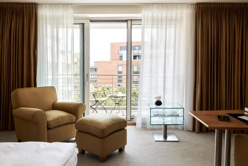Helles Hotelzimmer mit Sessel, Ottomane und Balkonblick auf moderne Gebäude