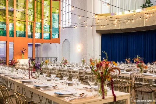 Festlich gedeckte Tafeln mit Blumen, Lichterketten und bunten Fenstern in hellen Saal.