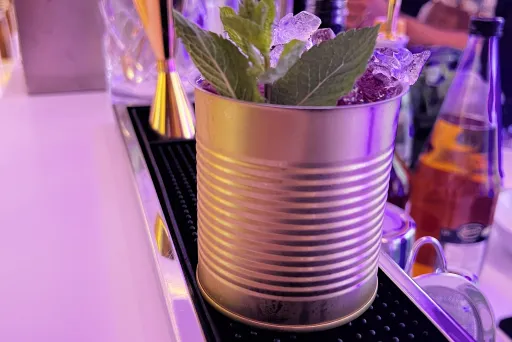 Cocktail in goldener Dose mit Eis und Minze auf einer Bar, lila Beleuchtung.