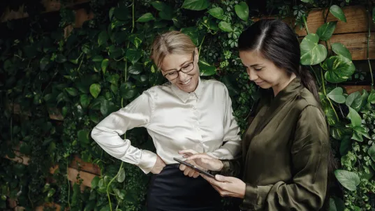 Zwei Frauen betrachten zusammen ein Tablet vor einer grünen Pflanzenwand.