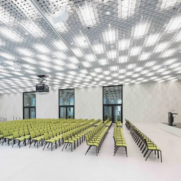 Moderner Konferenzsaal mit grünen Stühlen, großer Leinwand und futuristischer Decke.