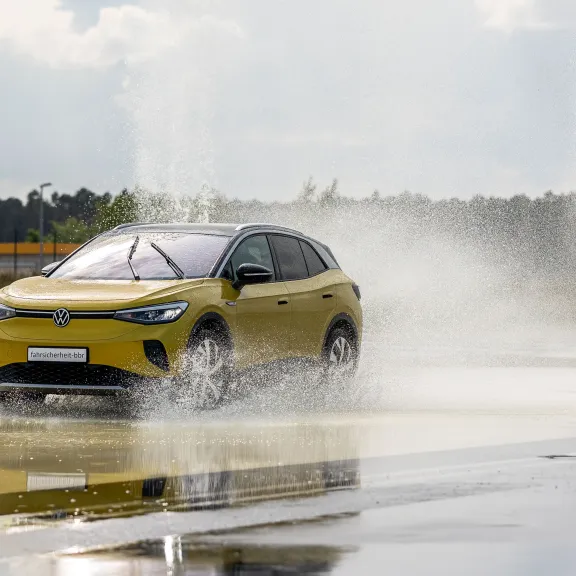 Gelbes Auto fährt durch große Wasserpfütze auf nasser Straße, Wasser spritzt hoch.