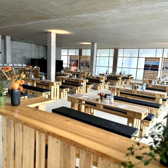 Großer, heller Raum mit Holzbänken und Tischen, modern dekoriert mit Blumen.