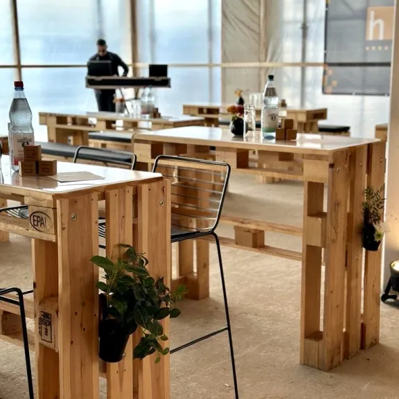 Helle Cafeteria mit Holztischen, Stühlen und Wasserflaschen, im Hintergrund eine Person.