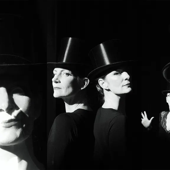 Schwarz-weiß-Foto von drei Frauen mit Zylindern, lächelnd und dramatisch beleuchtet.