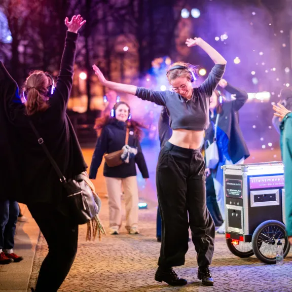 Teilnehmende mit leuchtenden Funkkopfhörern tanzen bei einer Silent Disco Tour im nächtlichen Stadtraum; im Hintergrund ein Technik-Cargo-Bike.