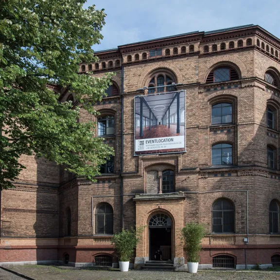 Backsteingebäude mit großem Werbebanner am Eingang, davor zwei Pflanzen.