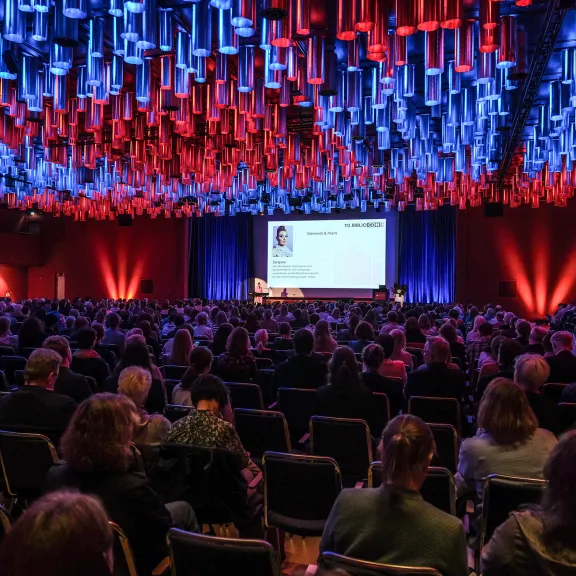 Großer Konferenzsaal mit vielen Zuschauern, Bühne vorne, bunte Deckenbeleuchtung.
