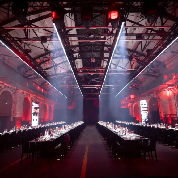 Langer Festsaal mit zwei gedeckten Tafeln, rote Beleuchtung, industrielle Atmosphäre.
