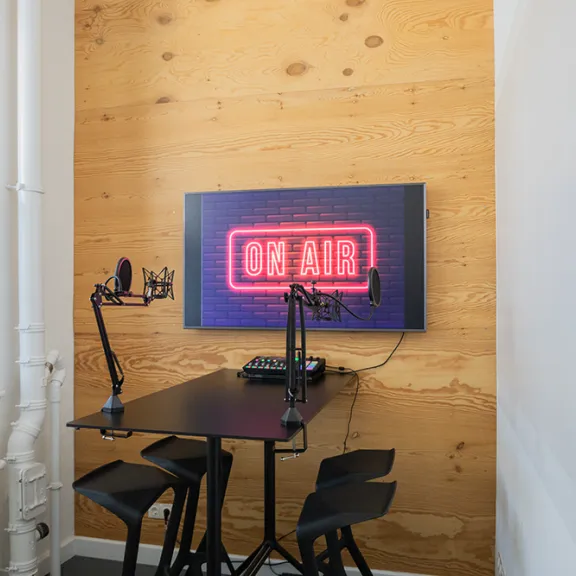 Kleines Podcast-Studio mit Tisch, Mikrofonen und leuchtendem "On Air"-Schild an der Wand.