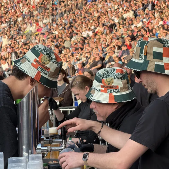 Drei Männer mit bunten Hüten schenken Getränke bei einem Festival aus, Menschenmenge im Hintergrund.