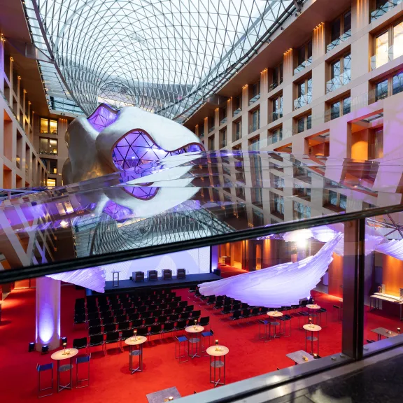 Modernes Atrium mit Glasdach, rotem Teppich und beleuchteter Eventfläche, Stuhlreihen und Tischen.