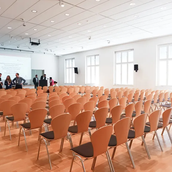 Heller Konferenzraum mit leeren Stuhlreihen und wenigen Menschen vorne am Rednerpult.