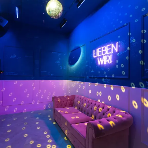 Bunte Lounge mit Disco-Kugel, pinkem Sofa und Neon-Schriftzug an der Wand.