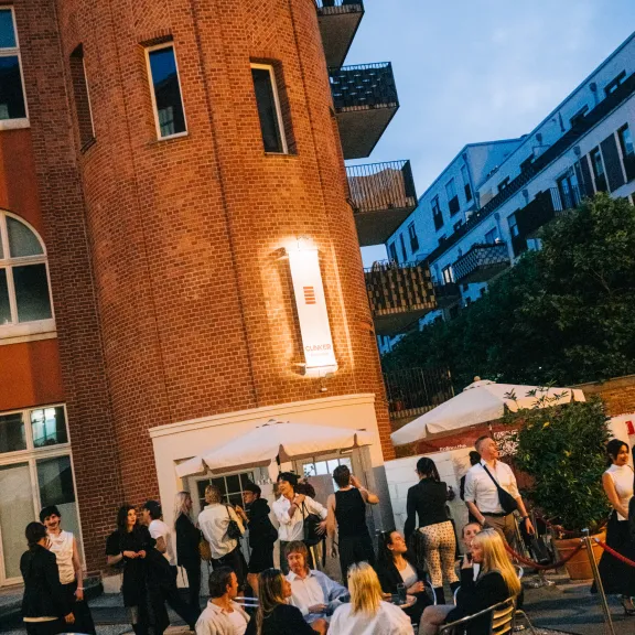Menschen sitzen und stehen abends vor einem Backsteingebäude mit Terrasse und Sonnenschirmen.