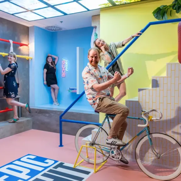 Drei lachende Erwachsene in farbenfrohem, verspieltem Raum mit Fahrrad und buntem Design.
