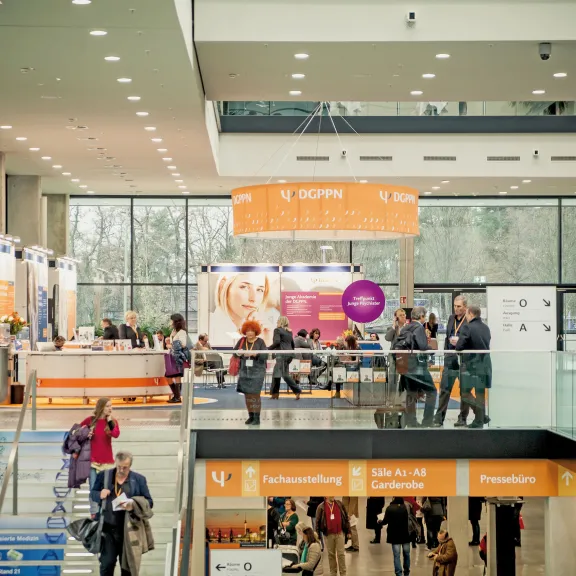 Menschen in einer modernen Messehalle mit Informationsständen und orangefarbenen Bannern.