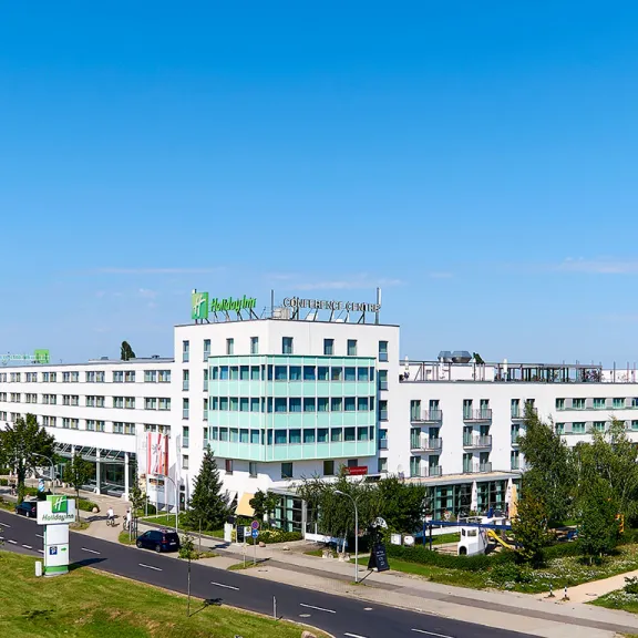 Modernes, weißes Hotelgebäude mit grünem Holiday Inn Logo und umliegender Begrünung.