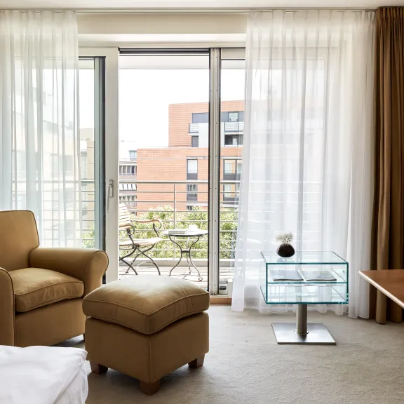 Helles Hotelzimmer mit Sessel, Ottomane und Balkonblick auf moderne Gebäude