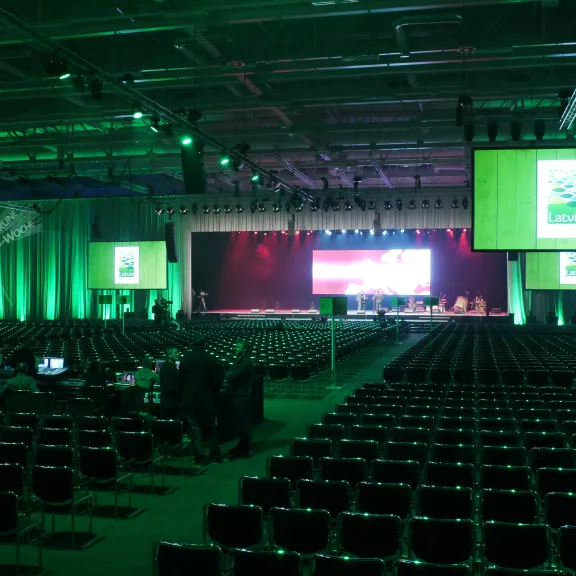 Großer Konferenzsaal mit grüner Beleuchtung, leeren Stuhlreihen und Leinwänden.