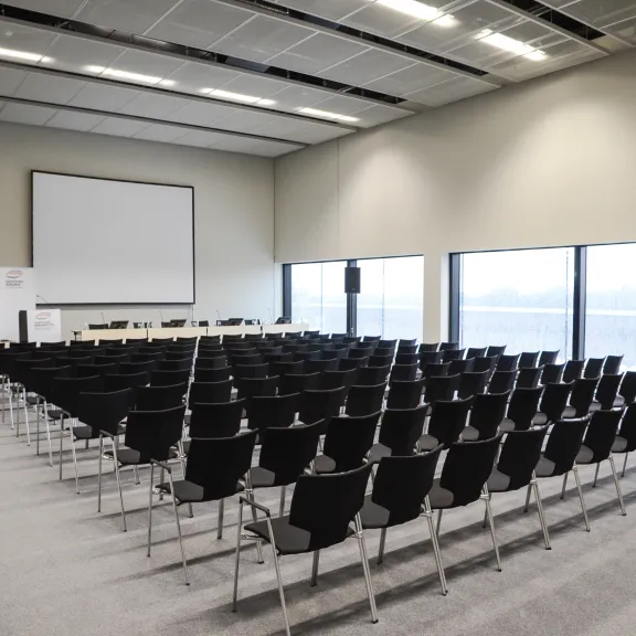 Leerer Konferenzraum mit schwarzen Stühlen, großem Fenster und Projektionsleinwand.