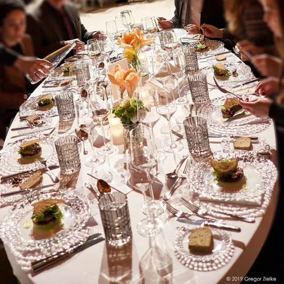 Festlich gedeckter Tisch mit Gläsern, Tellern und Blumen, mehrere Personen sitzen darum.