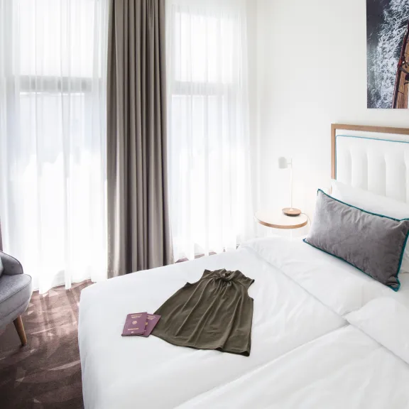Modernes Hotelzimmer mit Doppelbett, Kleid und Buch auf dem Bett, helle Vorhänge am Fenster.