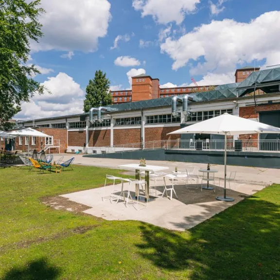 Garten mit Sitzgruppen und weißen Sonnenschirmen vor Backsteingebäude bei Sonnenschein.