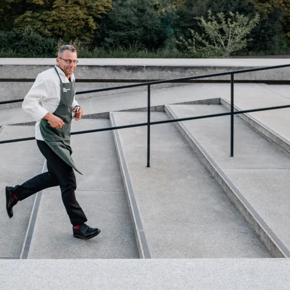 Mann mit Schürze läuft draußen eine breite Treppe hoch.