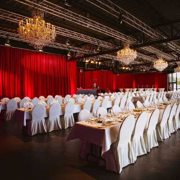 Eleganter Bankettsaal mit runden Tischen, weißen Stühlen und roten Vorhängen.