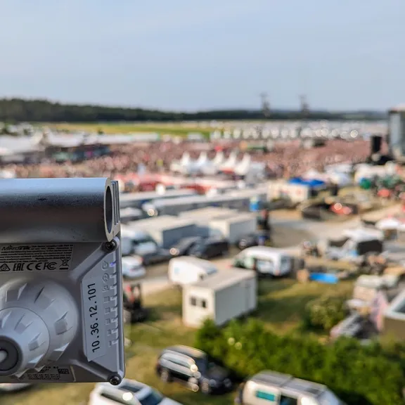 Überwachungskamera im Vordergrund, Festivalgelände mit Menschen im Hintergrund.