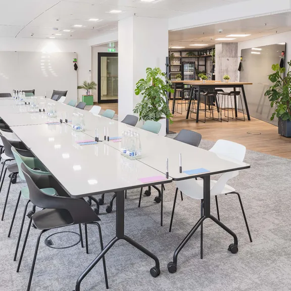 Heller Konferenzraum mit langem Tisch, vielen Stühlen und Pflanzen, moderne Einrichtung.
