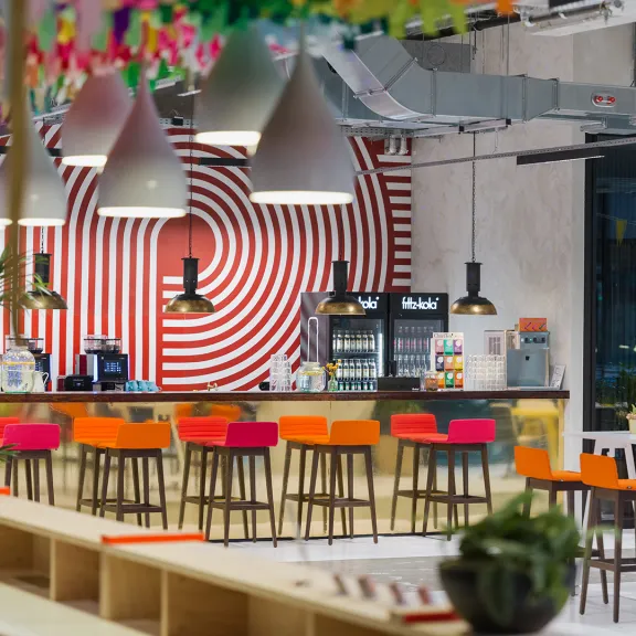 Moderne Cafeteria mit bunten Stühlen, Bar und auffälliger rot-weißer Wandkunst.