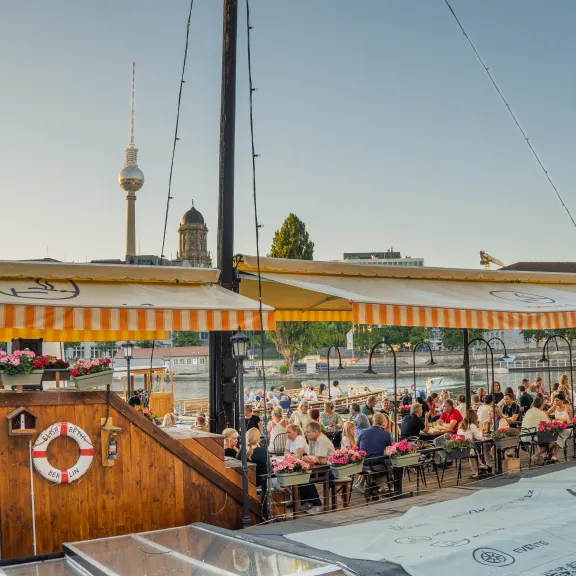 Sonnige Terrasse von Spree Fumée, Markisen spenden Schatten, zahlreiche Gäste genießen das Ambiente bei strahlendem Wetter.