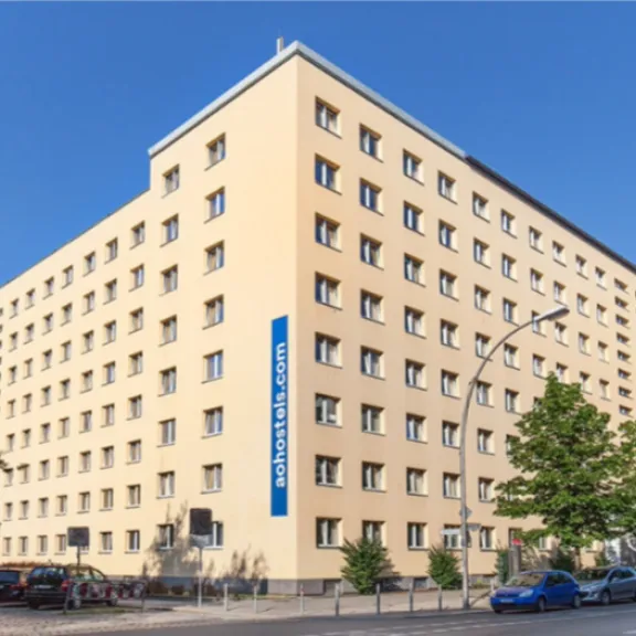 Großes, beiges Hotelgebäude mit mehreren Stockwerken an einer Straßenecke bei Sonnenschein.