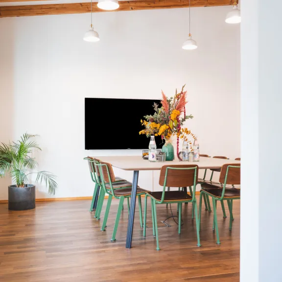 Sichtbar ist ein modern eingerichteter Meetingraum mit einem zentralen Tisch und 10 Stühlen. Der Raum ist hochwertig eingerichet mit einem Bildschirm und viel Helligkeit.