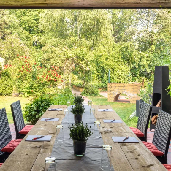 Langer Holztisch mit Stühlen auf Terrasse, Blick in grünen Garten.