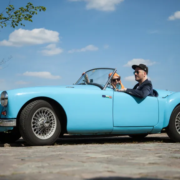Mann mit Hund fährt bei Sonnenschein in einem blauen Cabrio über Kopfsteinpflaster.