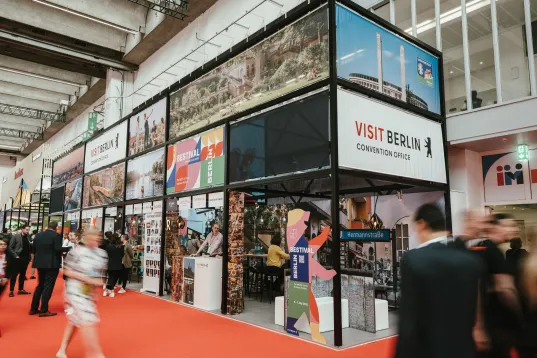 Besuchende gehen durch die Messehalle an einem Stand von visitBerlin mit Bildern der Stadt vorbei.