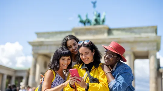 Eine Gruppe von vier Frauen steht vor dem Brandenburger Tor und schaut lächelnd auf ein Smartphone, das eine der Frauen in der Hand hält