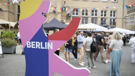Buntes Berlin-Bären-Schild auf Hof mit Menschen und Wimpeln beim BESTIVAL Festival. 