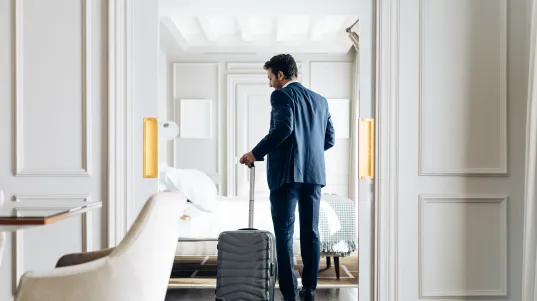Mann im Anzug betritt mit Rollkoffer ein elegantes Hotelzimmer.
