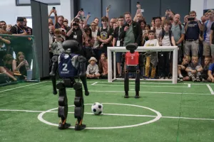 Zwei humanoide Roboter spielen Fußball auf einem Kunstrasenfeld, Zuschauer im Hintergrund.